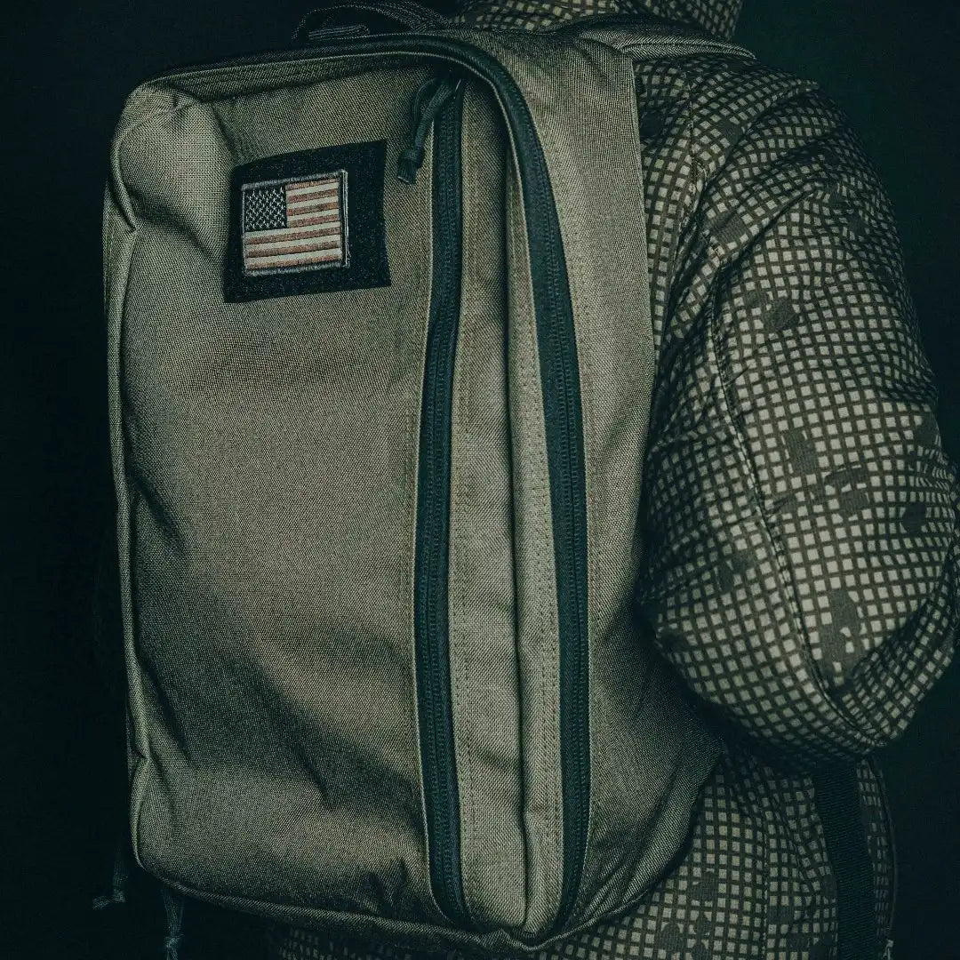 Person in a patterned jacket wearing the olive green Recycled Firefighter 24 Hour Backpack with a US flag patch, side view