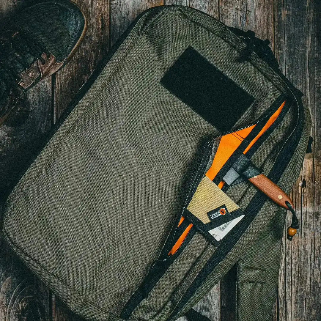 Top-down view of an olive green Recycled Firefighter 24 Hour Backpack on a wooden surface with an orange-lined open pocket holding small items