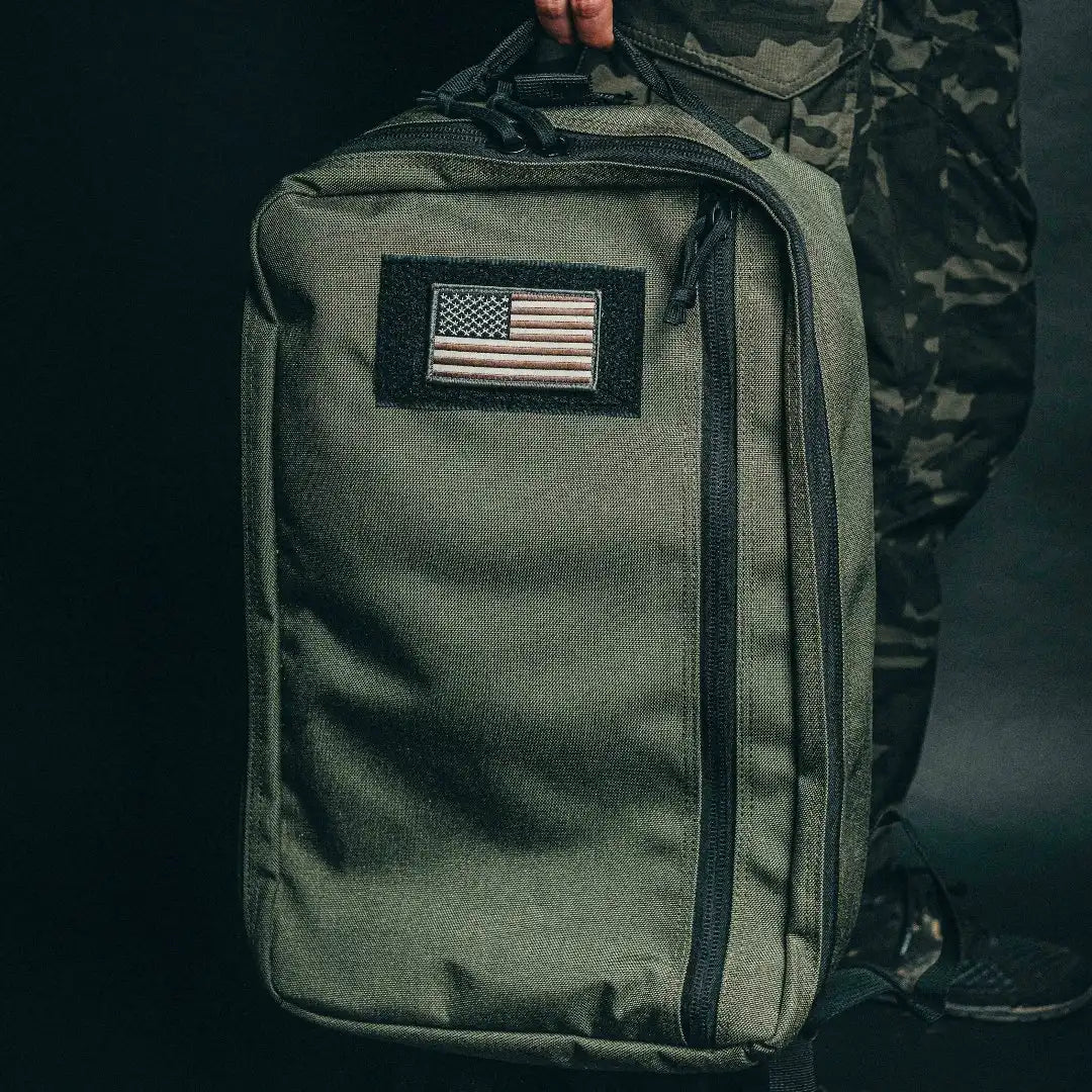 Person holding the olive green Recycled Firefighter 24 Hour Backpack with a US flag patch by its top handle