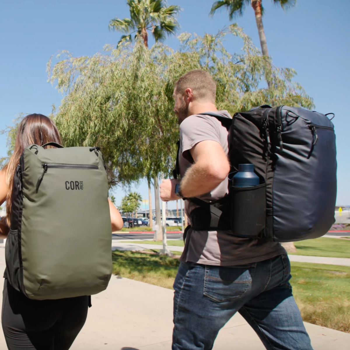 Two hikers walking outdoors wearing Island Hopper backpacks, one olive green and one dark navy
