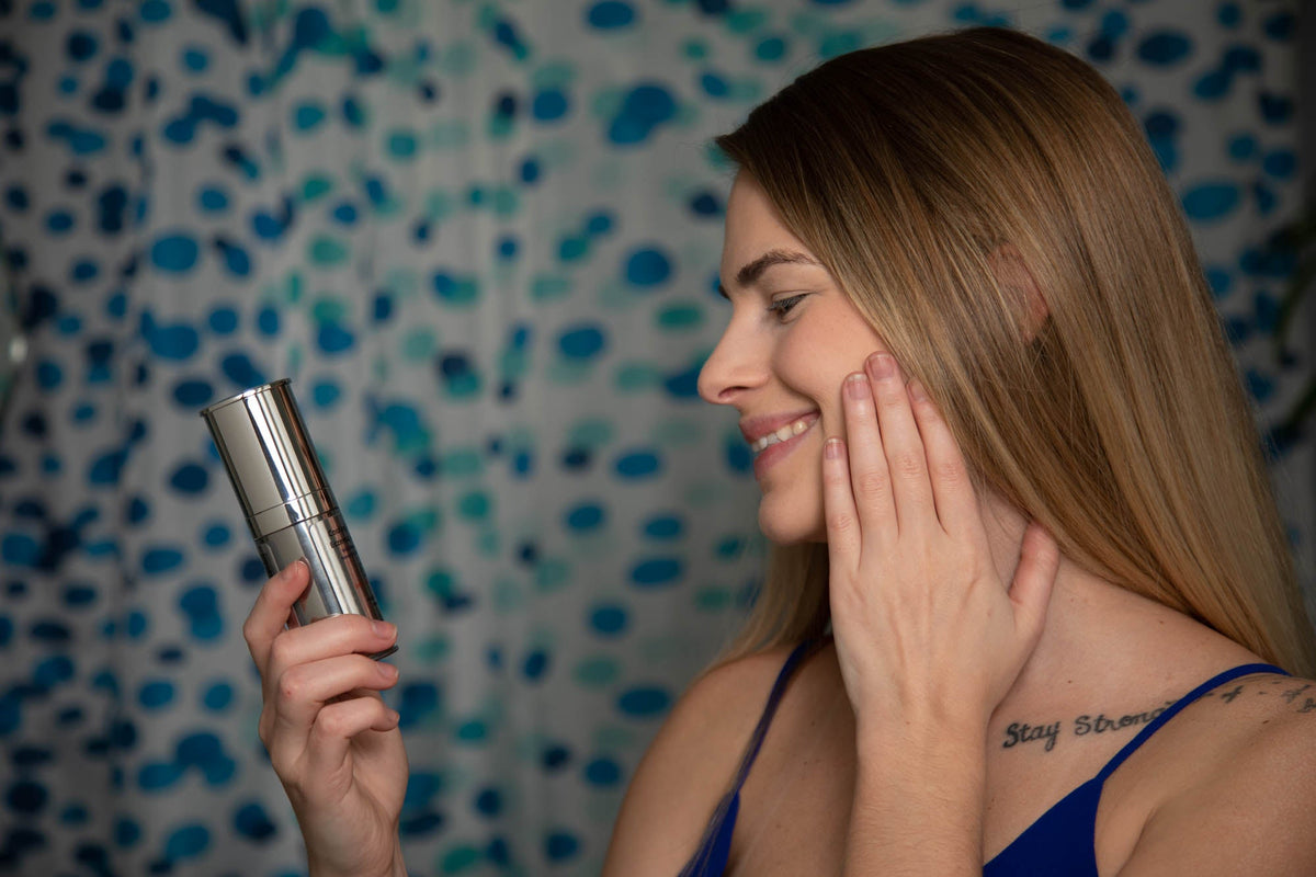 Woman touching her cheek while holding the EpiLynx moisturizer bottle in front of a blue patterned wall