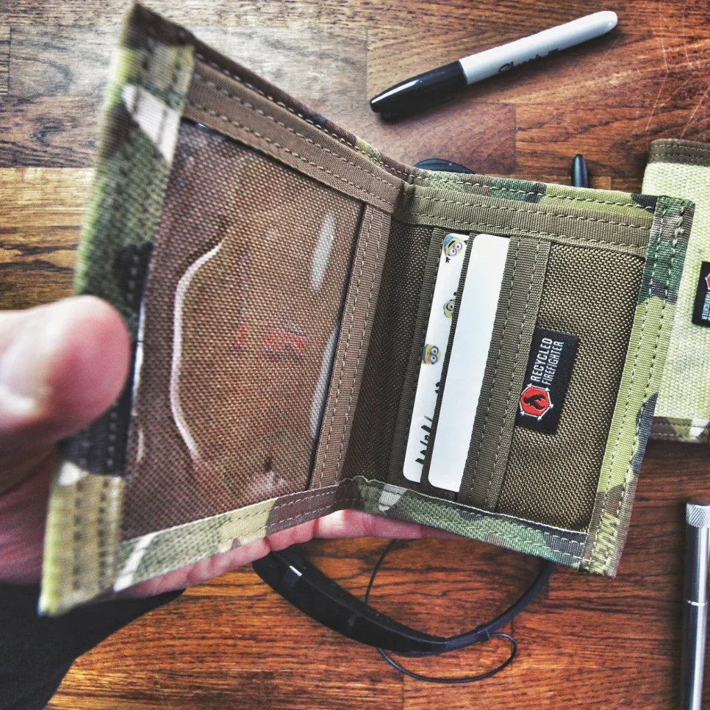 Hand holding the open camouflage wallet with cards in the slots on a wooden table beside a pen