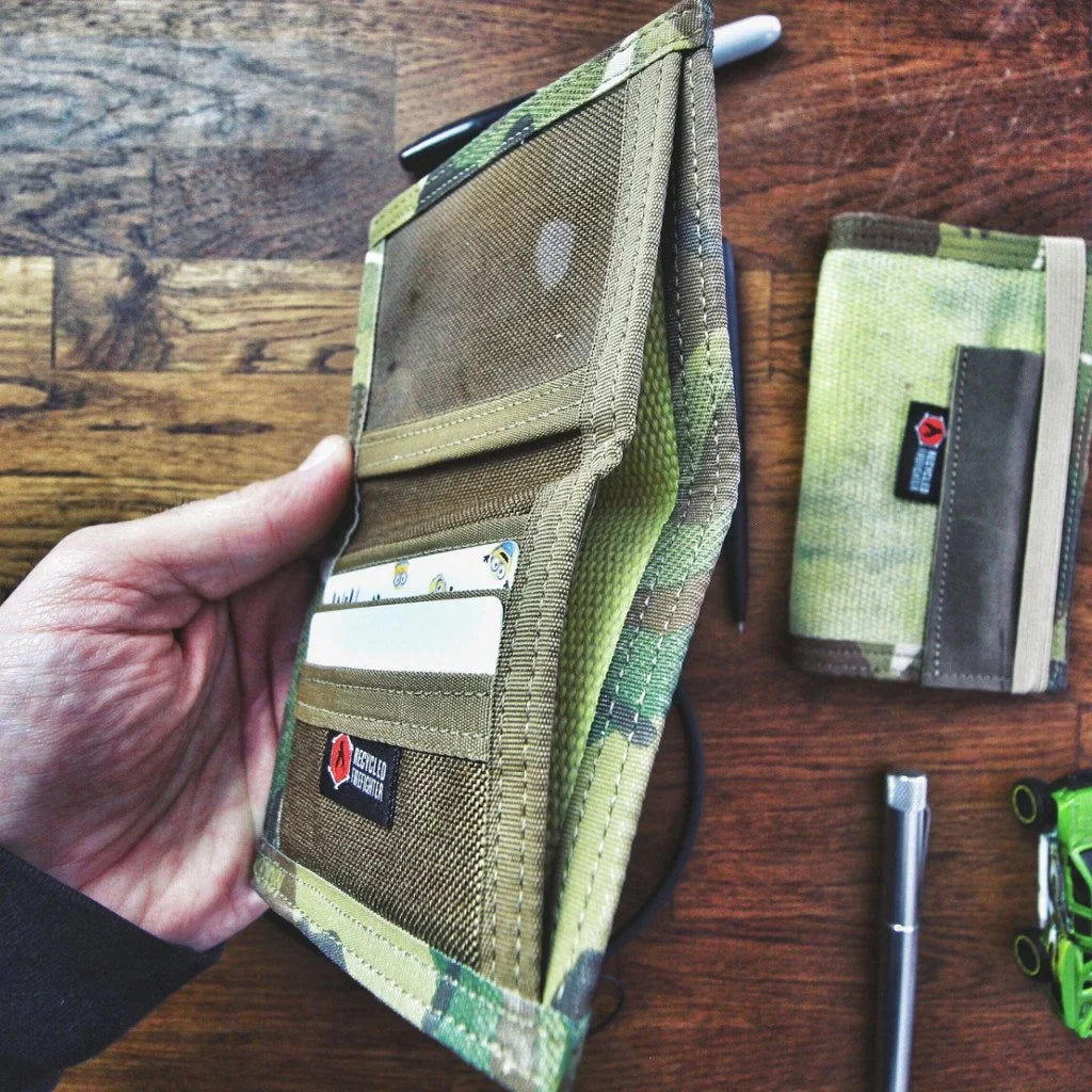 Open camouflage wallet held next to a second wallet on a wooden table with a pen