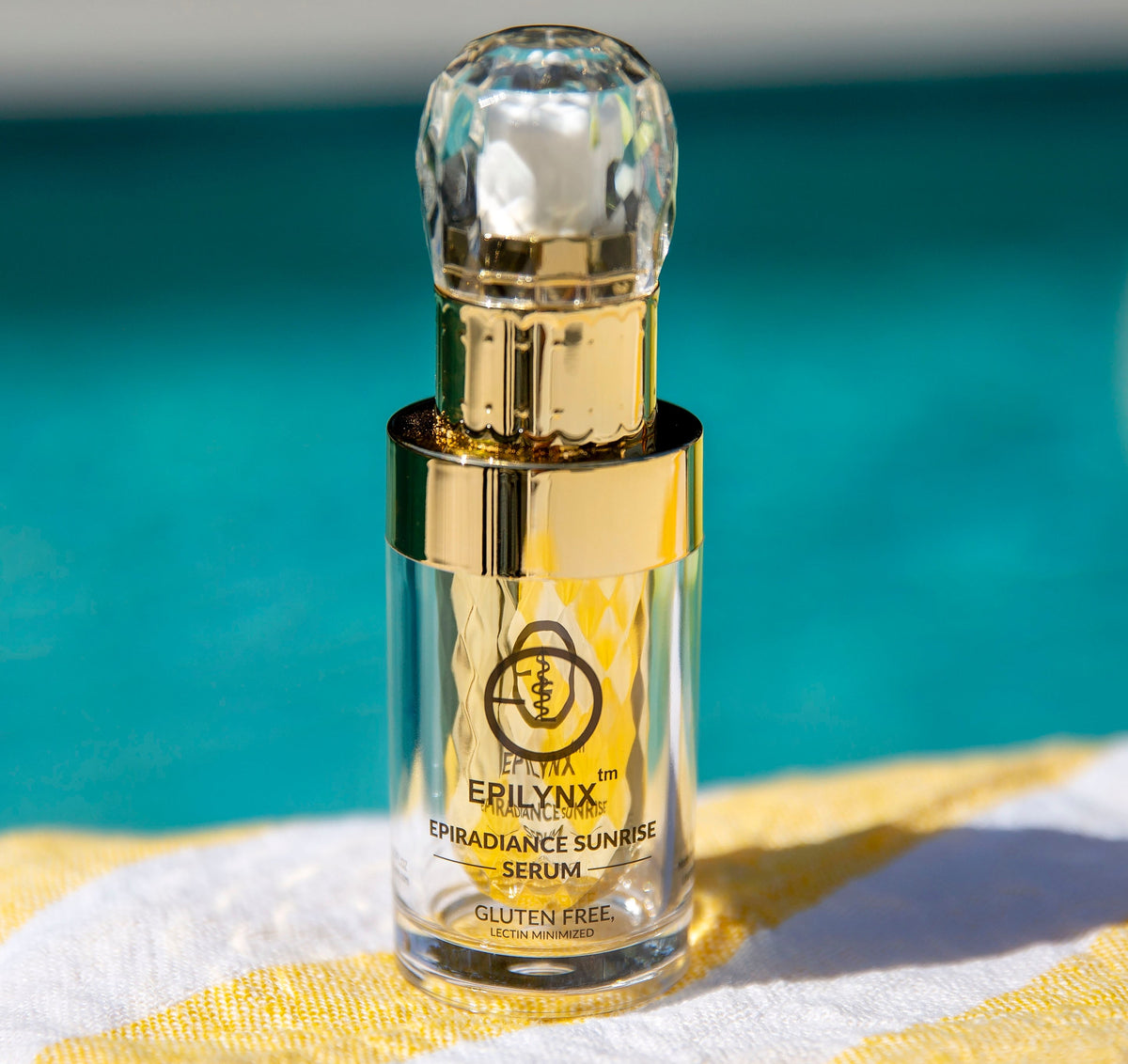 Gold EpiLynx Sunrise serum dropper bottle on a folded yellow and white towel beside a turquoise pool