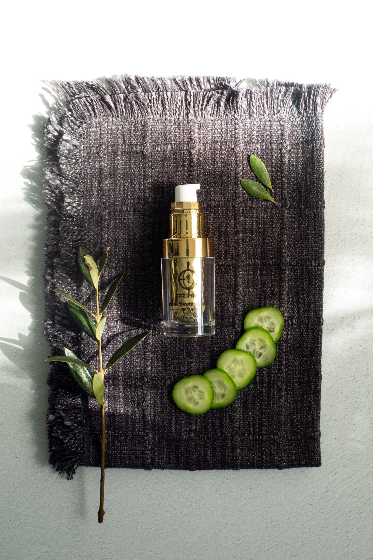 Gold serum bottle on a dark cloth with cucumber slices and a sprig of leaves