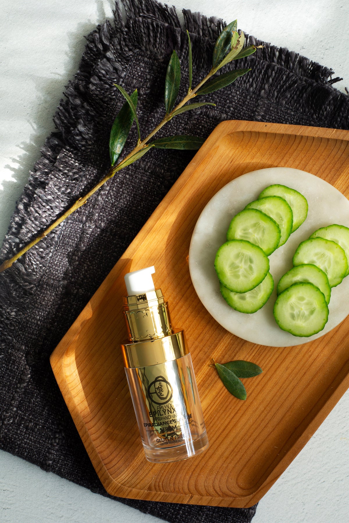 Gold serum bottle on a wooden tray with cucumber slices and olive leaves, top view