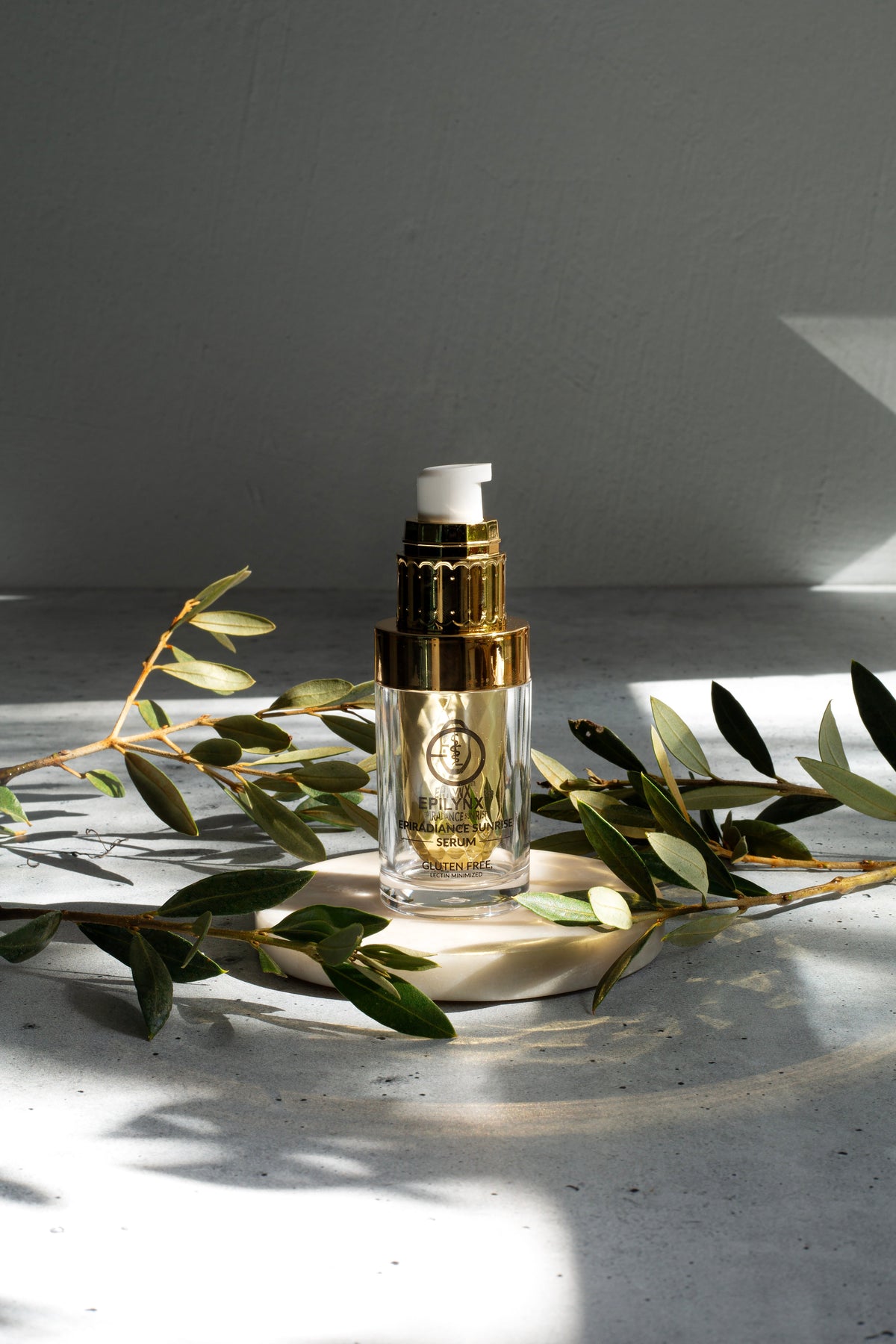 Gold serum bottle surrounded by olive branches in soft sunlight