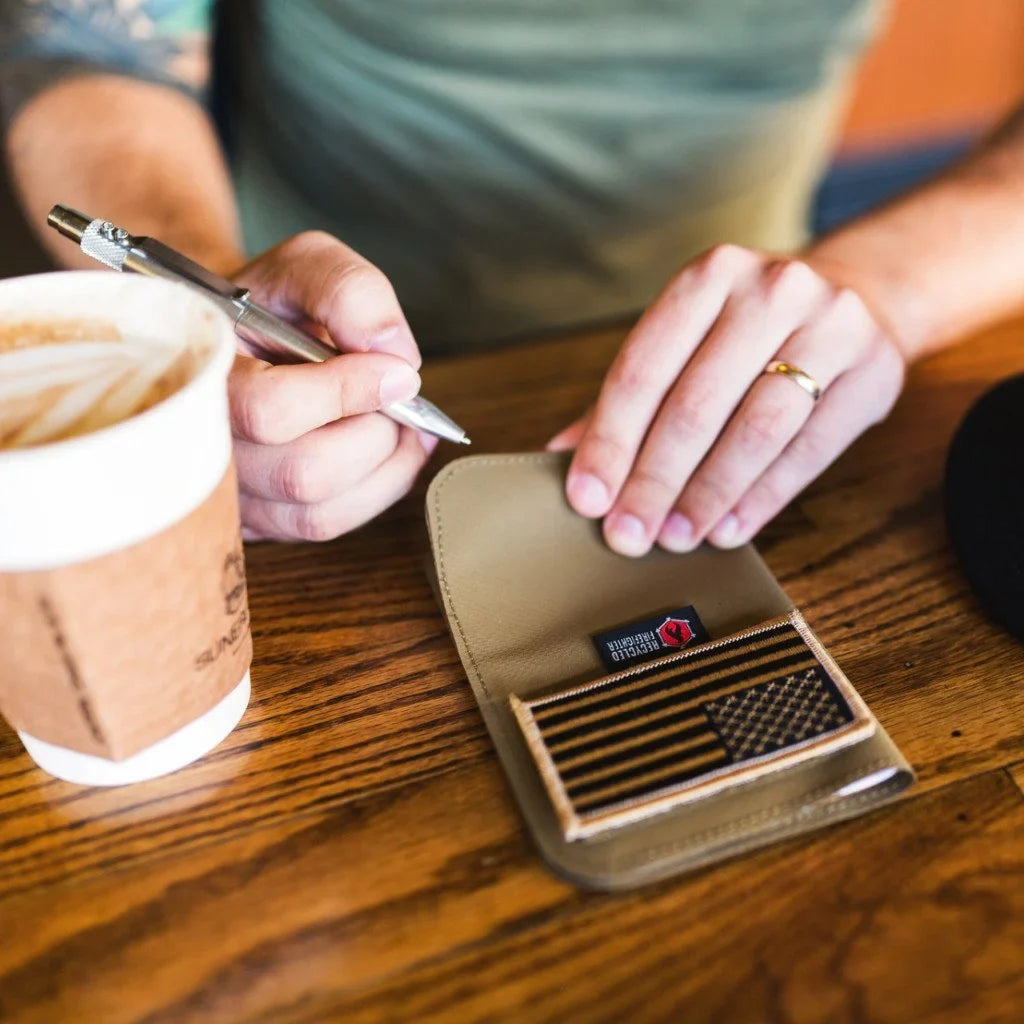 Hand holding a pen over the tan Field Notes Wallet on a wooden table beside a coffee cup
