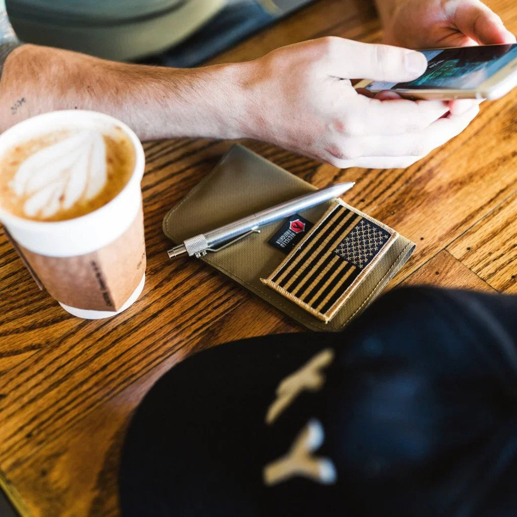 Field Notes Wallet with a flag patch and a pen on a wooden table beside a latte and a person holding a phone