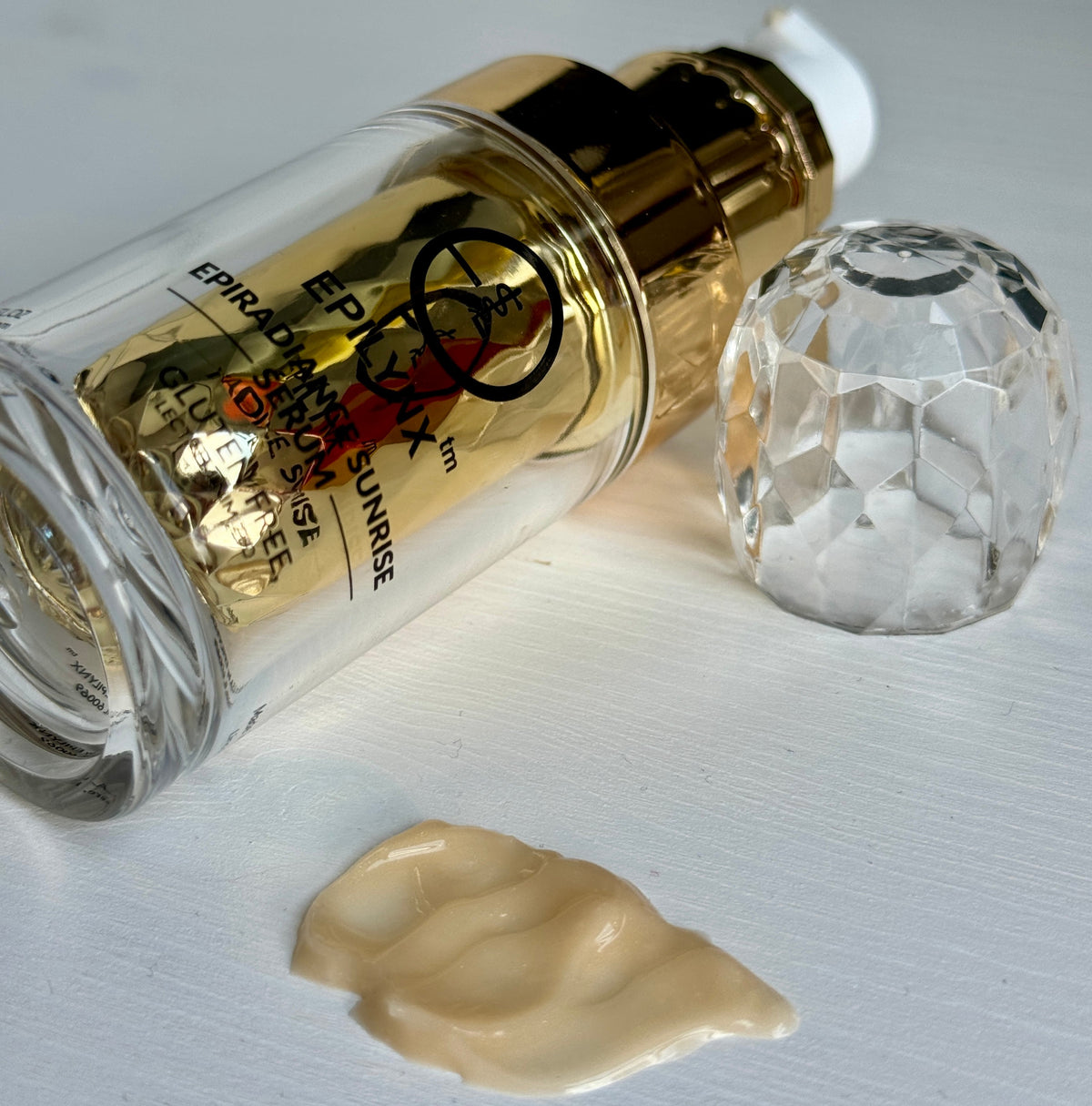 Gold serum bottle lying on its side beside a clear crystal-style cap and a smear of cream