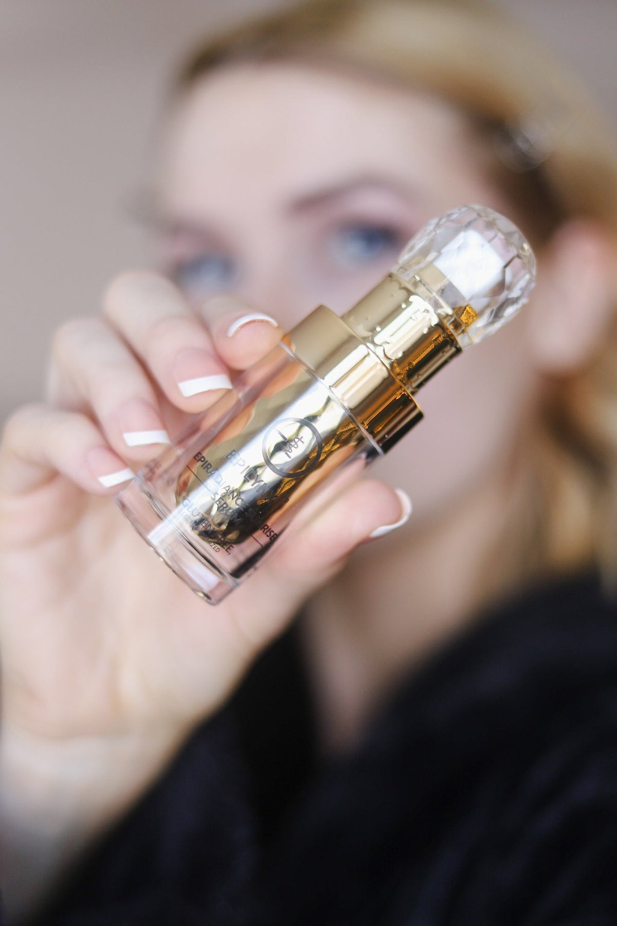 Woman in a black robe holding the gold serum bottle toward the camera
