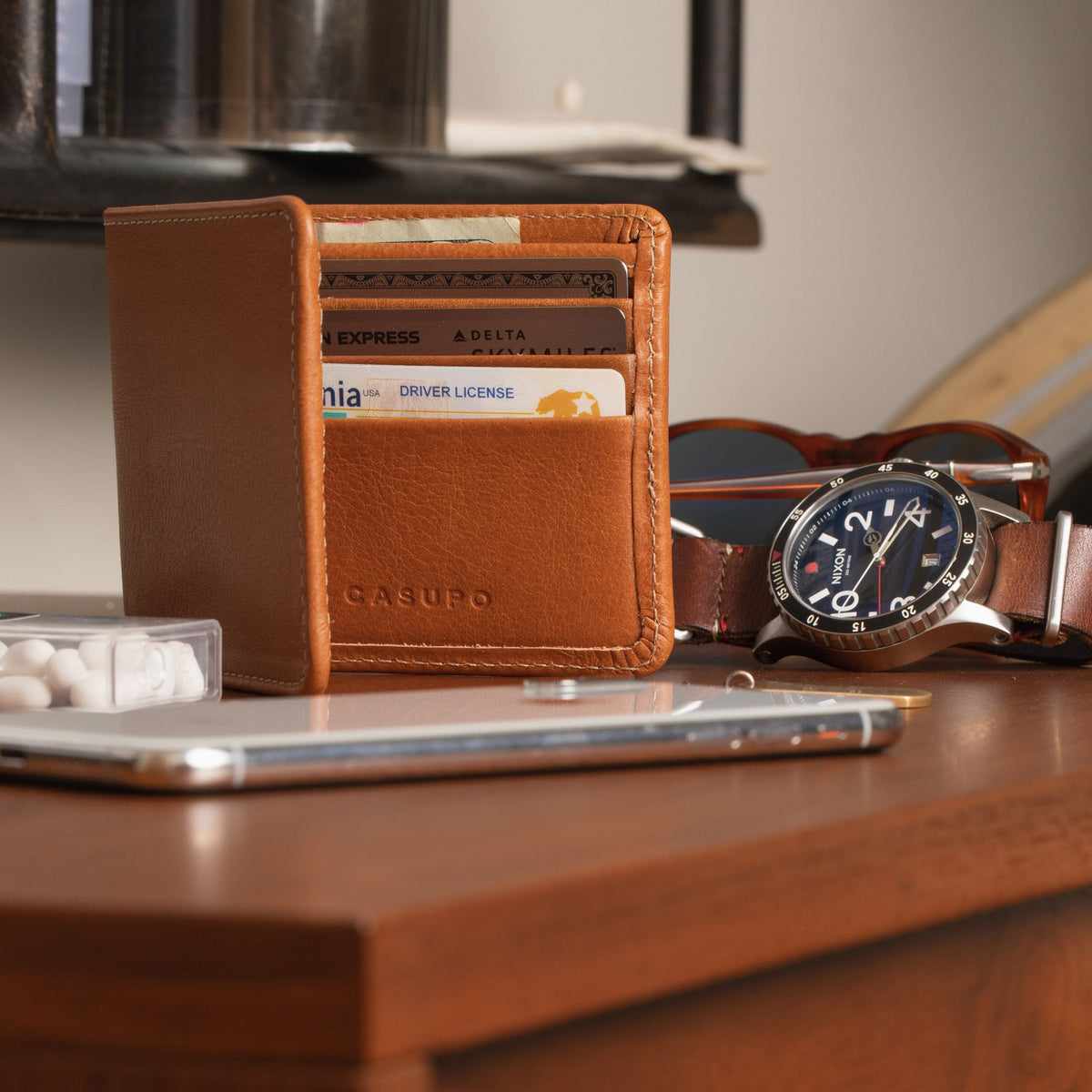 Tan leather billfold standing open on a wooden desk beside a watch and a phone