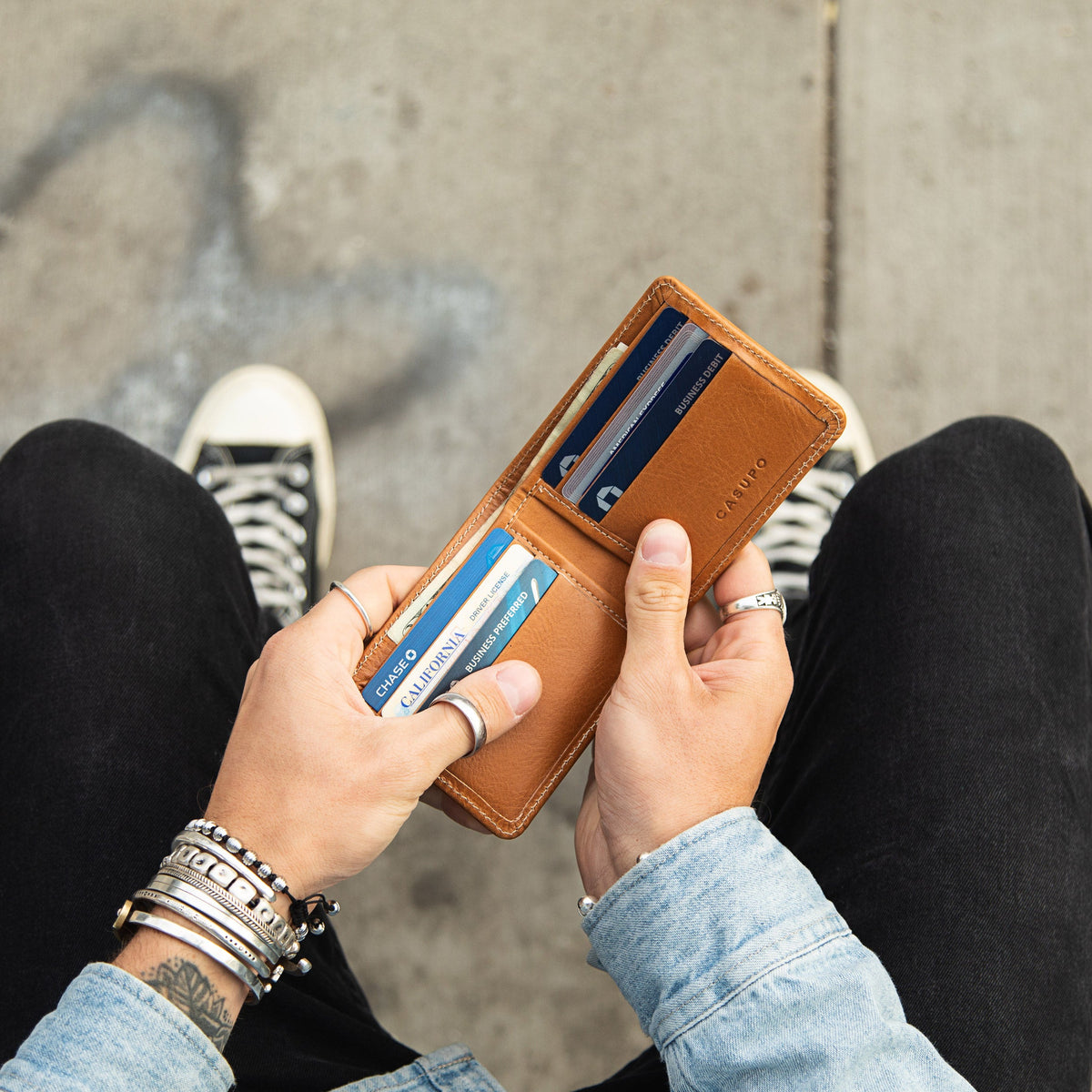 Hands holding the open tan leather billfold with cards in the slots, seen from above with sneakers on the pavement