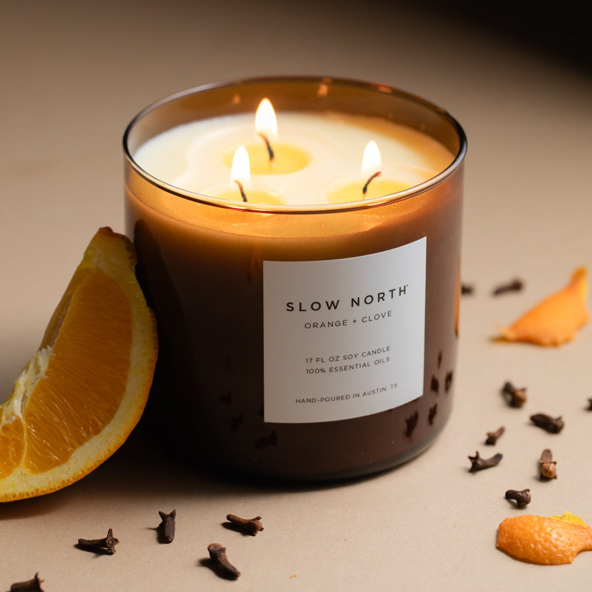 Lit three-wick candle in an amber jar beside an orange slice and scattered cloves