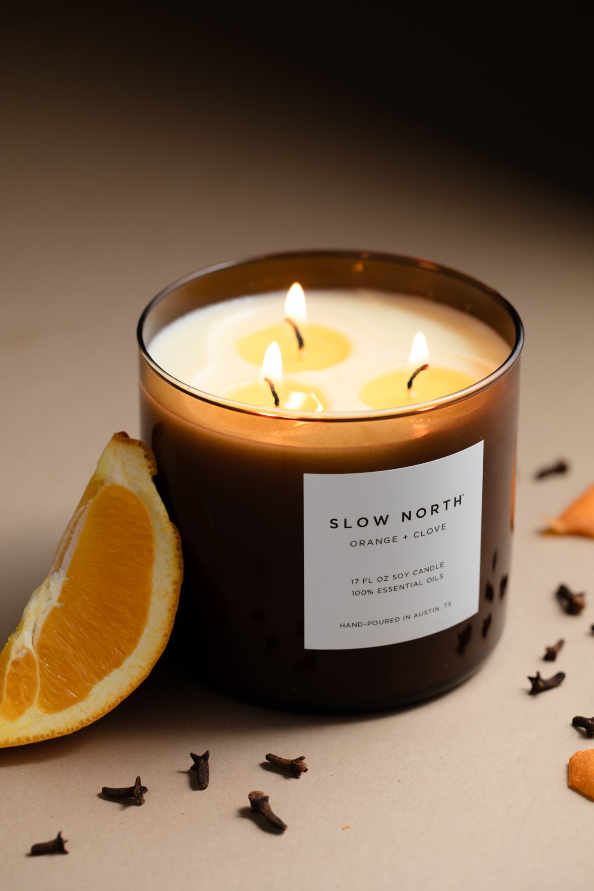 Lit candle in an amber jar beside an orange slice and cloves against a dark background