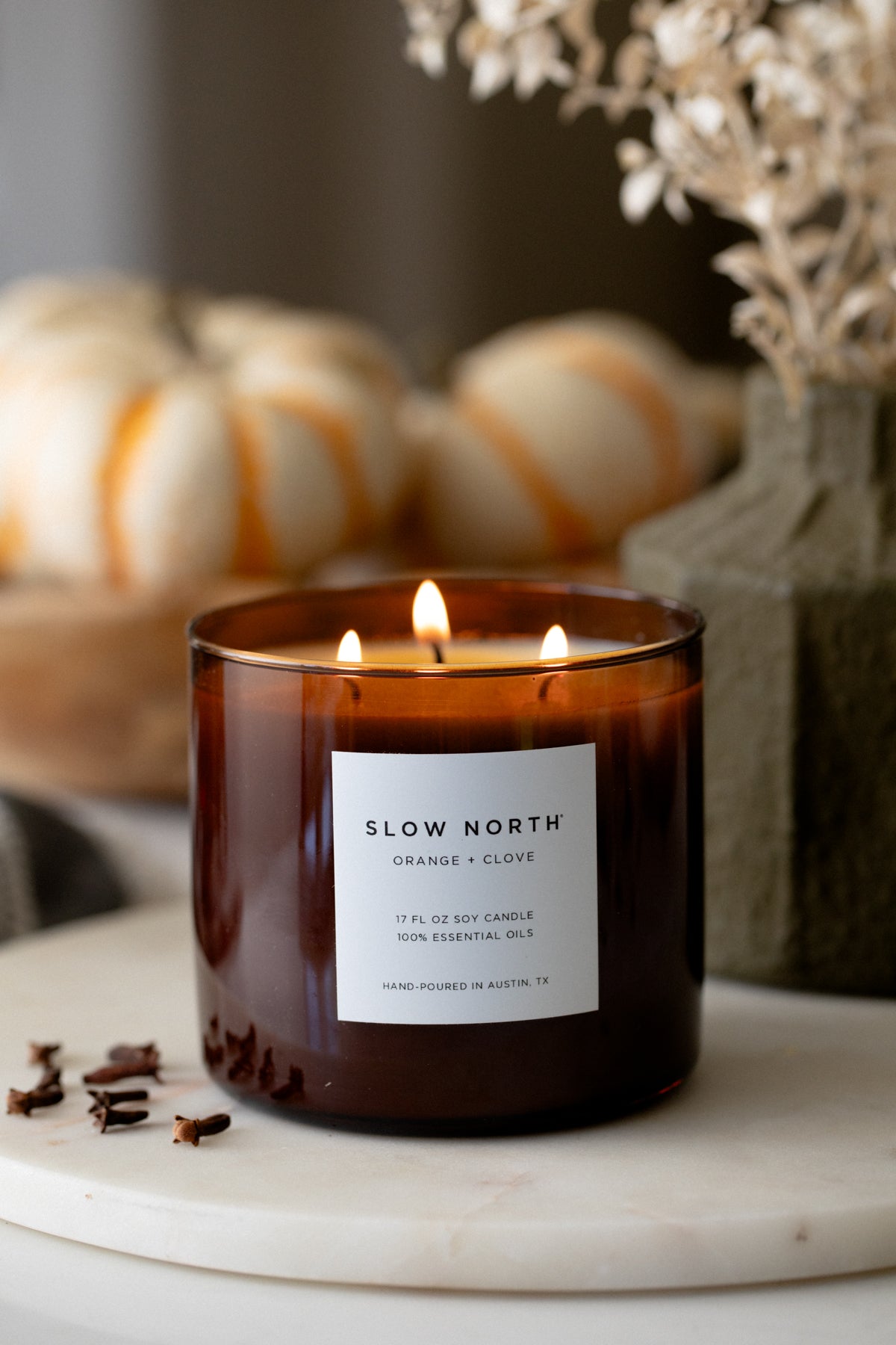 Lit candle in an amber jar on a marble tray beside small pumpkins and dried flowers