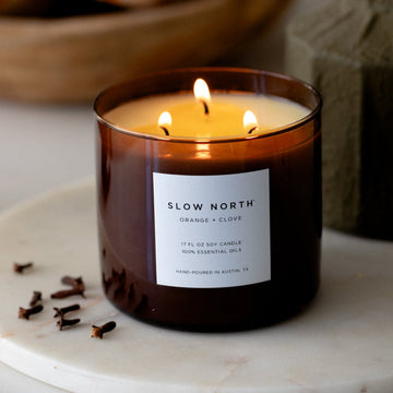Slow North Orange + Clove three-wick candle in an amber glass jar with a white label, lit on a marble tray beside cloves