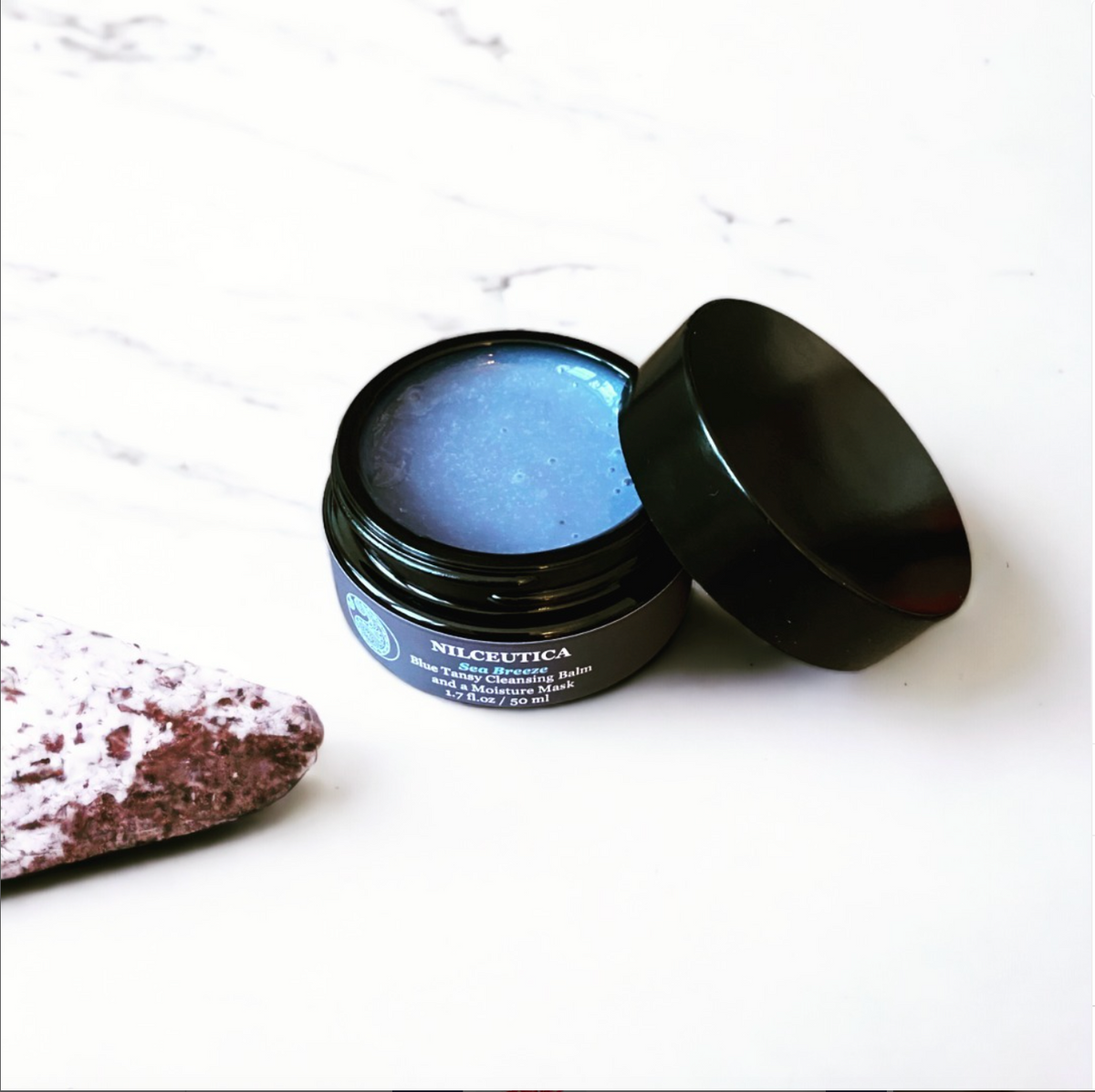 Open round jar of blue balm beside its black lid on a white marble surface