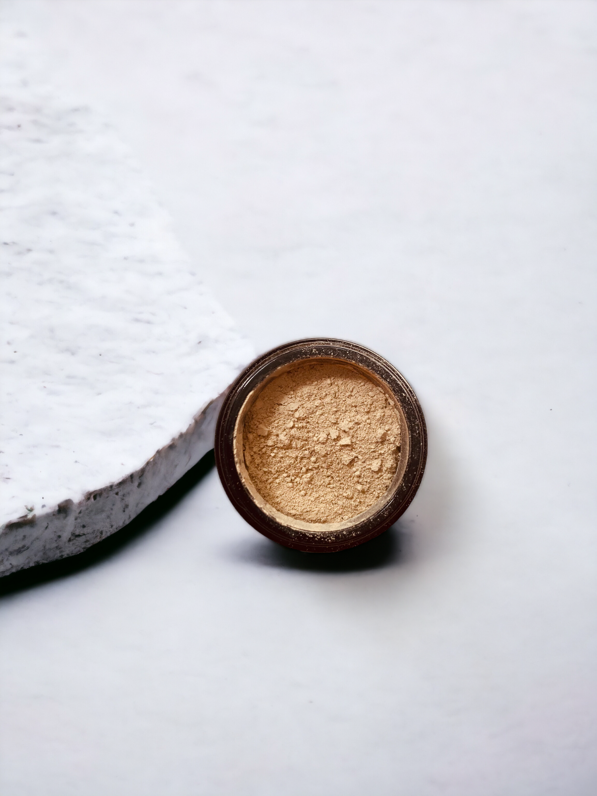 Small dark container of beige powder beside its lid on a white background