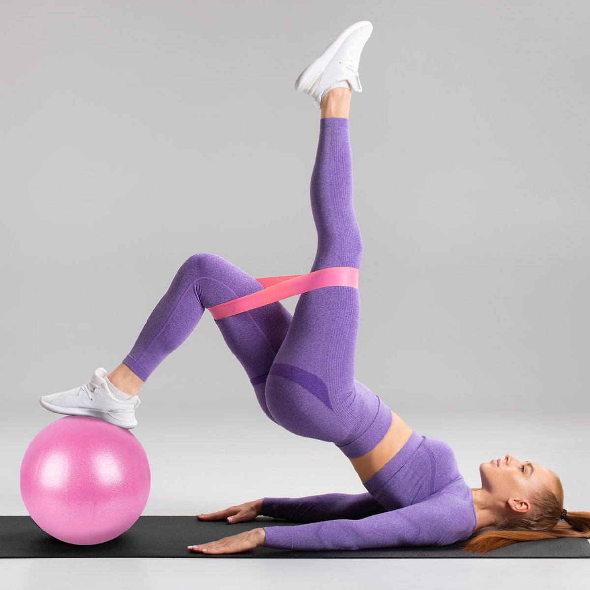 Woman in purple workout wear doing a bridge with a pink resistance band and a pink exercise ball