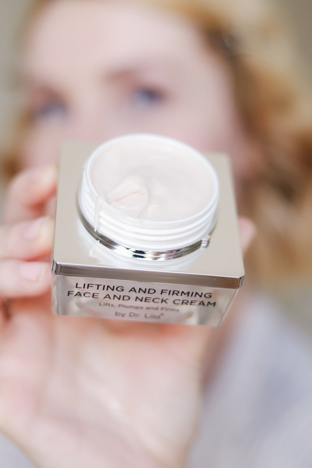 Hand holding the open cream jar toward the camera, blurred background
