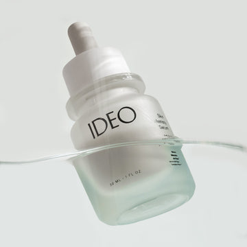 IDEO Skin Memory Serum dropper bottle lying in shallow water on a light gray background