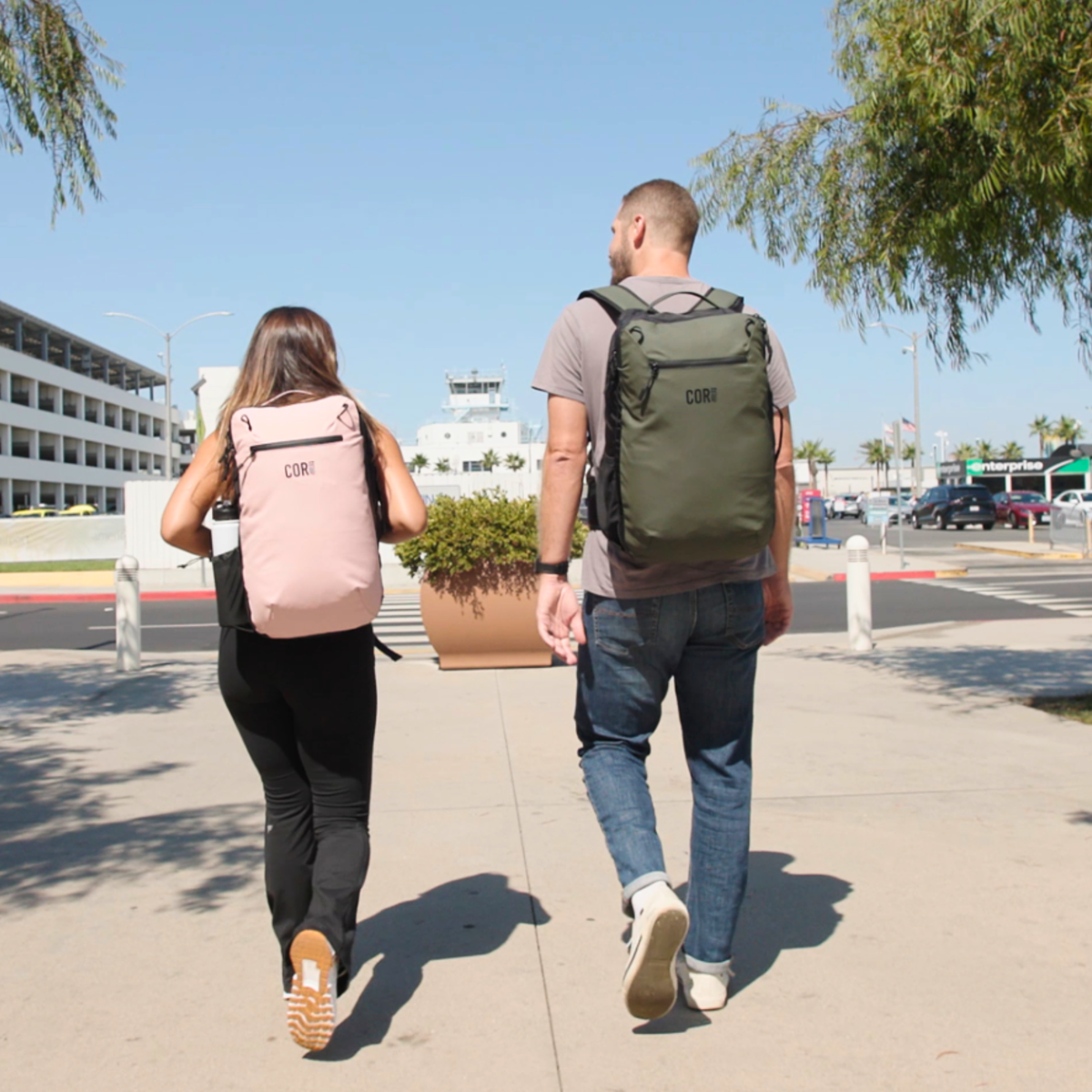 Two travelers seen from behind walking through an airport area, one with a pink backpack and one with an olive green backpack