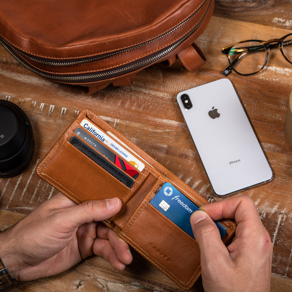 Hands pulling a blue card from the tan leather billfold on a wooden table beside a phone and glasses