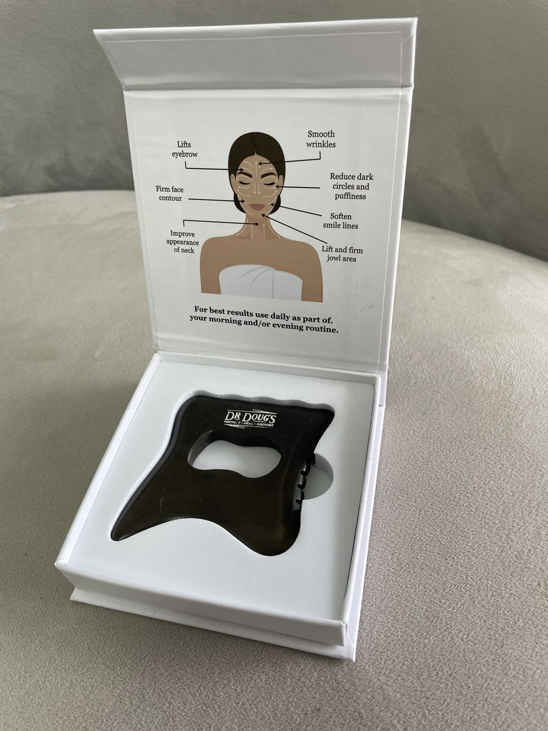 Open white box at an angle holding a small gua sha tool, with an instruction card showing a woman's face