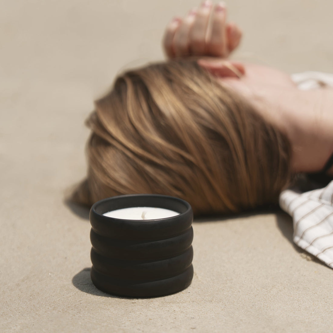 Black ribbed ceramic candle with a white wax top on a sandy surface beside a woman lying with her head turned away